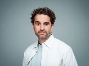 Steffen Boettcher Oliver Wnuk 207895 Ein Mann mit lockigem braunem Haar und Bart, der ein weißes Hemd über einem hellblauen T-Shirt trägt und vor einem schlichten grauen Hintergrund steht.